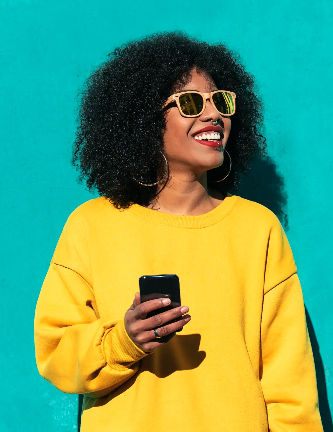 Vrouw met telefoon in een gele trui tegen een turquoise muur